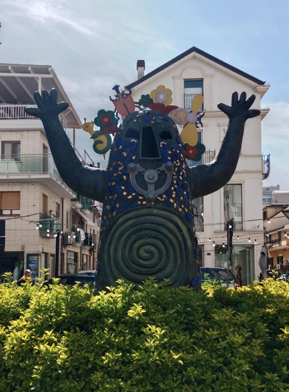 Un pupazzo gigante che dovrebbe far paura ma che invece è simpatico. È colorato di un blu scuro, ha una spirale sulla pancia e del pezzame di color oro e blu su tutto il corpo, un naso grande trapezoidale e una bocca meccanica fatta col pezzo di nonsoche. Da dietro la testa gli escono fiori, animali e insetti. Ha le braccia alzate e le mani aperte, nel gesto di mettere paura, uuuh. Di fronte al pupazzo c'è una siepe bassa, dietro di lui ci sono degli edifici e delle automobili.